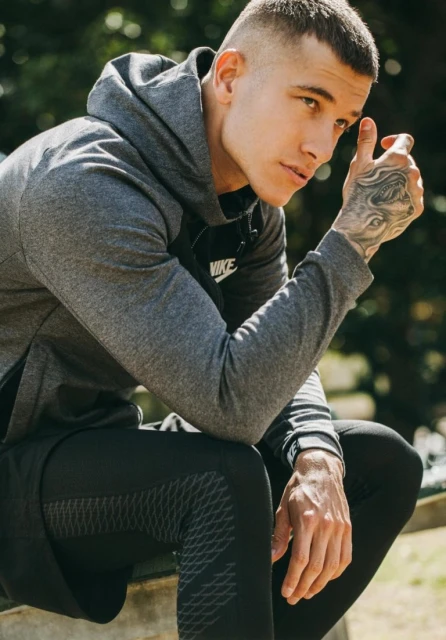 Athletic man in a grey hoodie and black athletic leggings sits outdoors in the sunlight, showcasing casual fitness fashion and style.