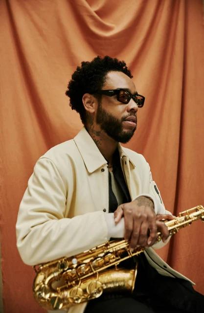 Adult male musician with a beard and glasses holds a gold saxophone in front of a draped orange background in a studio setting.