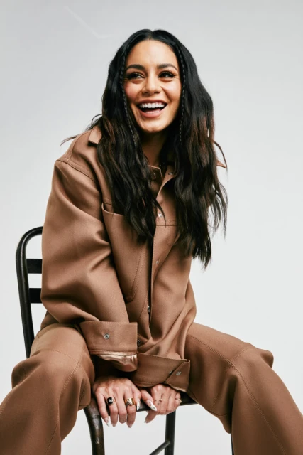 Vanessa Hudgens sits on a black chair in a photo studio wearing a beige jumpsuit with long, dark hair, smiling and looking at the camera.