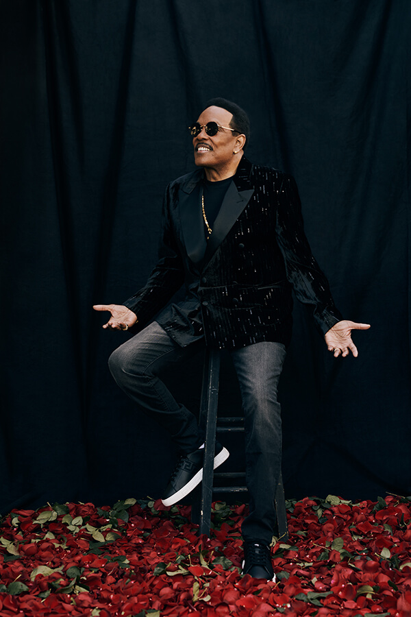 Portrait of a Black adult male with sunglasses, wearing a black velvet suit and black sneakers, sitting on a ladder on a floor of rose petals.