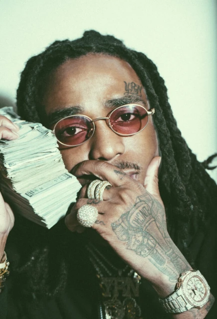 Portrait of rapper Quavo wearing glasses and jewelry, holding a stack of cash to his face in an indoor setting with fair lighting.