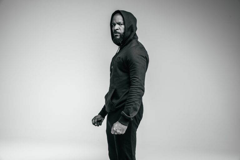 Black and white portrait of a black man wearing a dark hooded sweatshirt against a seamless studio background