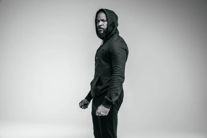 Black and white portrait of a black man wearing a dark hooded sweatshirt against a seamless studio background