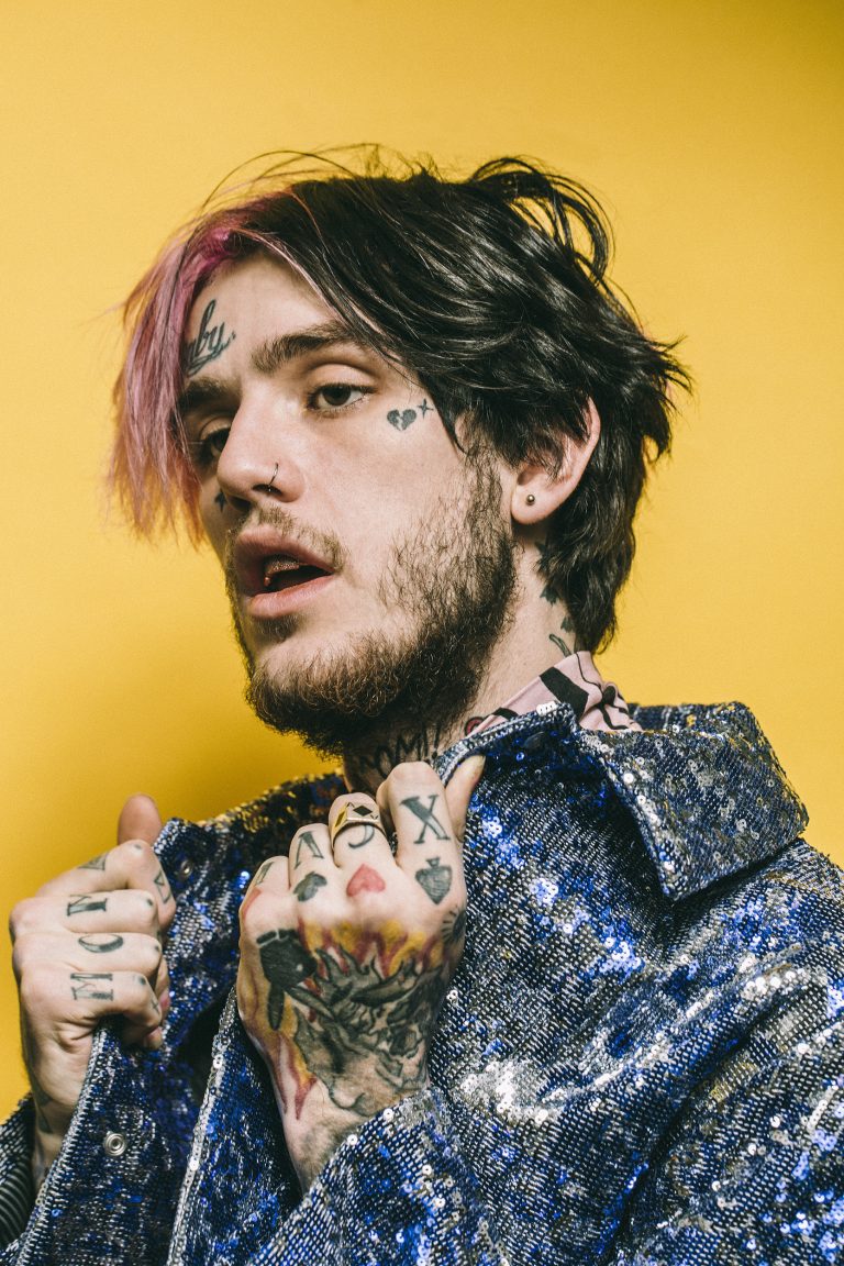 Portrait of a young adult male with facial tattoos wearing a sequin jacket against a yellow background. He has pink-tipped dark hair.
