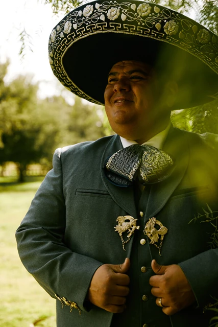 Portrait of a smiling adult male in a dark mariachi suit with gold trim and a sombrero, posing outdoors among green trees.