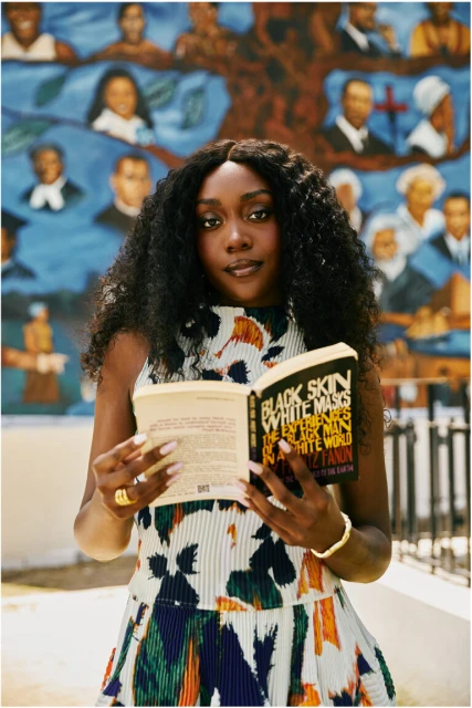 Portrait of a young Black woman reading 'Black Skin, White Masks' in front of a colorful mural, showcasing fashion and literary appreciation.