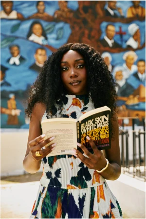 Portrait of a young Black woman reading 'Black Skin, White Masks' in front of a colorful mural, showcasing fashion and literary appreciation.