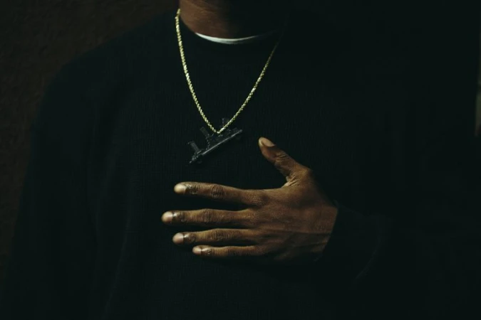 Close-up portrait of a man wearing a black sweater, gold chain, and gun pendant photographed by G L Askew Ii