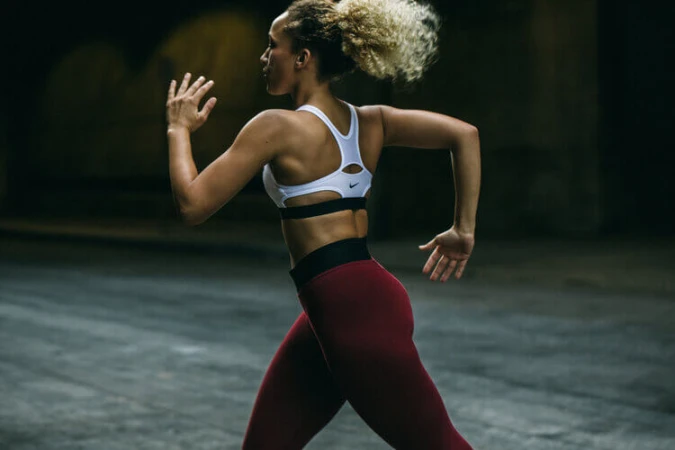 Athletic woman with curly blonde hair running in a white sports bra and dark red leggings against a dark blurred background.