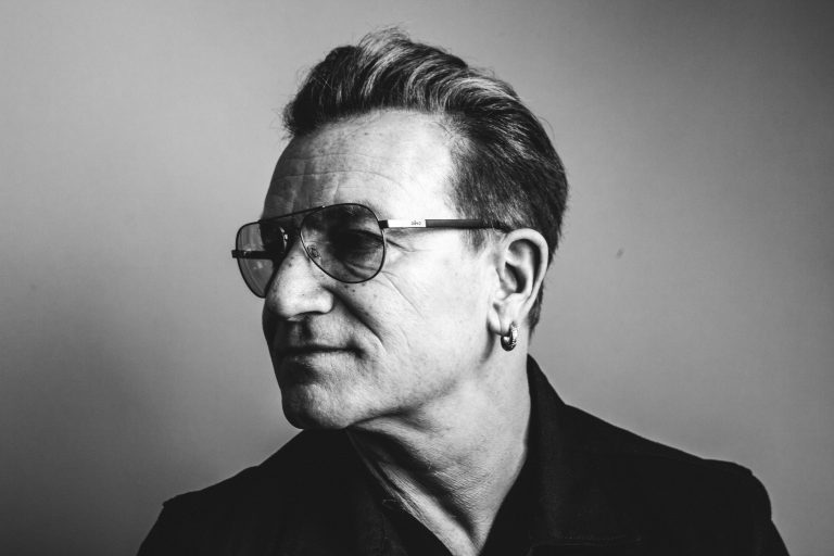 Black and white portrait of a mature adult male wearing aviator glasses and a dark jacket against a neutral grey backdrop.