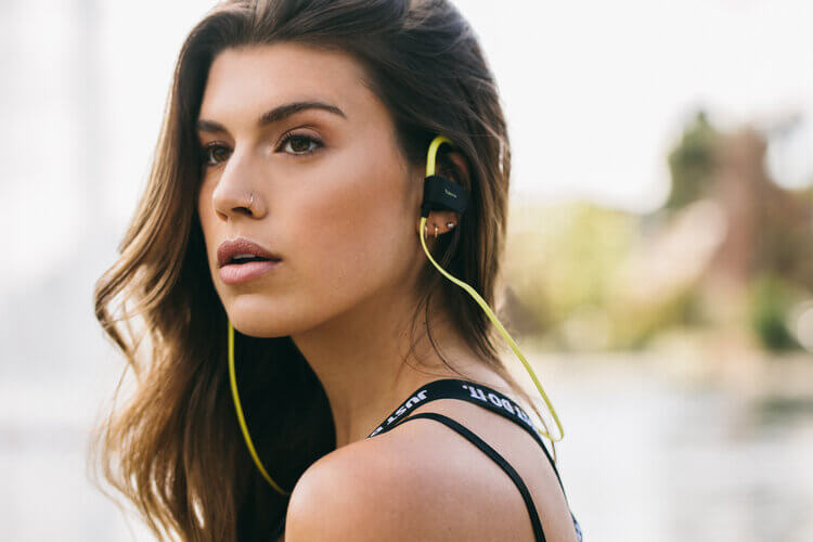 Attractive brunette woman wears wireless earbuds and a black sports bra with text. Outdoors, blurred water and trees in background.