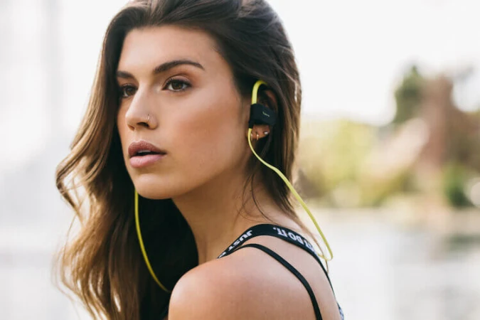 Attractive brunette woman wears wireless earbuds and a black sports bra with text. Outdoors, blurred water and trees in background.