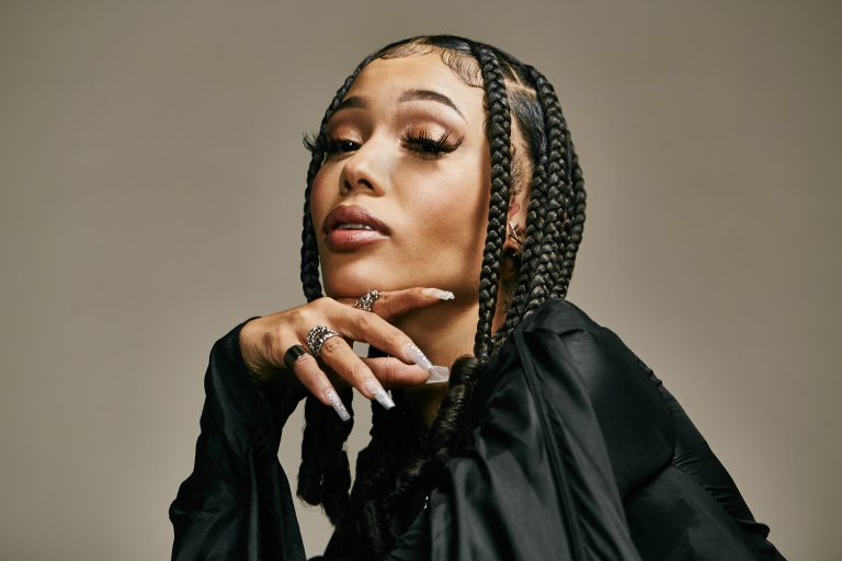 Close-up portrait of a woman with braided hair and several rings posing against a solid tan background in a photo studio.