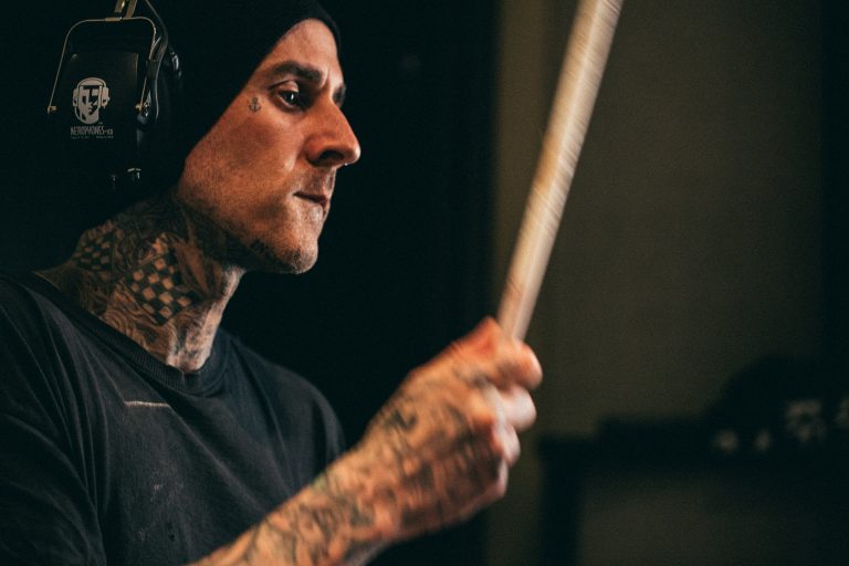 Close-up of a tattooed man with a beanie and headphones holding a drumstick in a music studio. The lighting is soft and diffused.