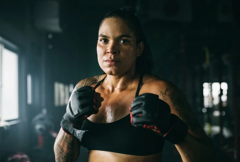 Determined female fighter in sports bra and gloves stands ready in a dimly lit gym, showcasing strength and focus.