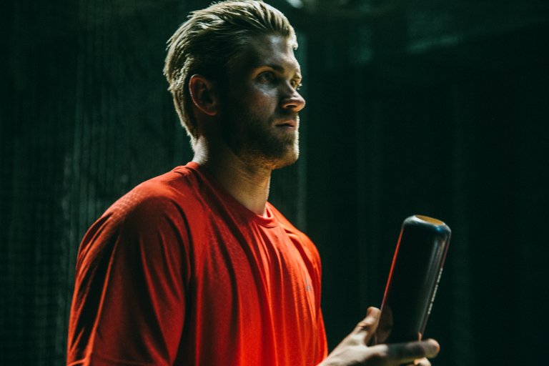 Adult male athlete in red shirt holding a baseball bat in a dimly lit indoor setting, close-up shot, profile view, intense mood.