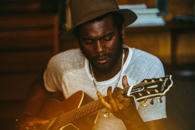 African American musician plays acoustic guitar indoors. The man wears a white shirt and fedora while strumming guitar strings.