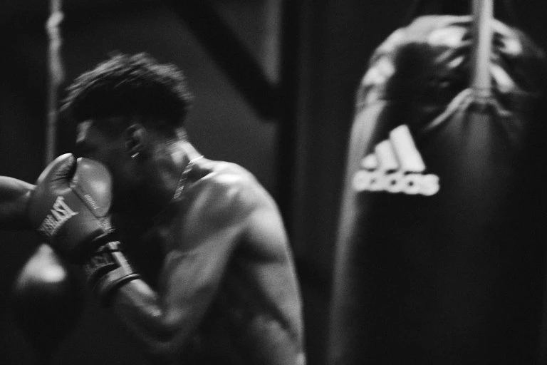 Monochromatic shot of a muscular adult male boxing with gloves at a gym; with a punching bag and minimal lighting, creates a moody atmosphere.