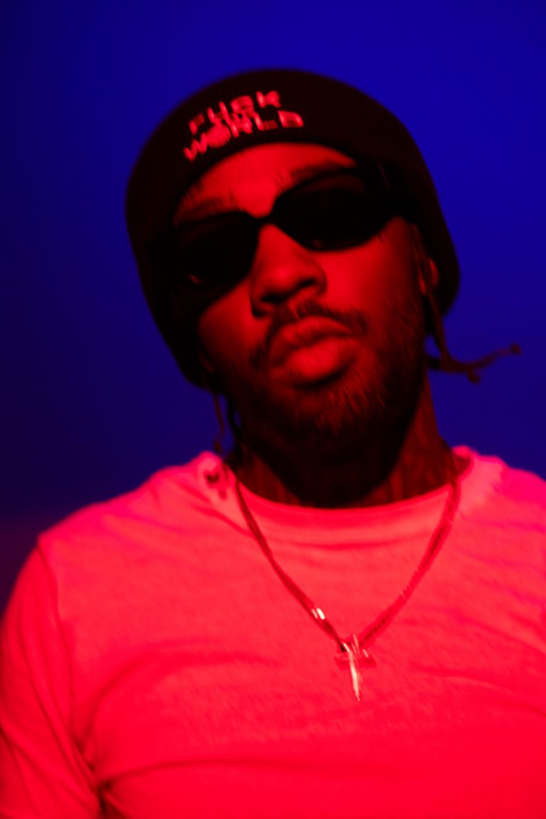 Portrait of a man wearing sunglasses and a black beanie with red text against a blue background with pink and red lighting.
