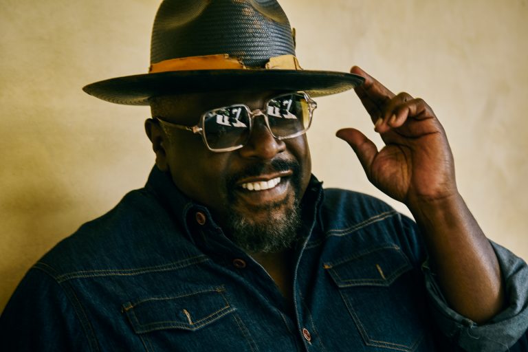Portrait of an African American man wearing a hat and glasses, looking at the camera with a neutral expression indoors.
