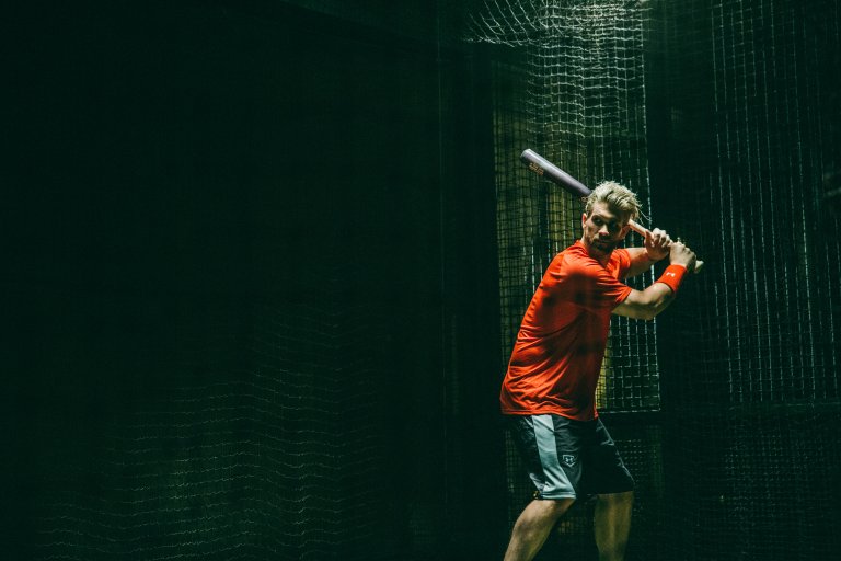 Adult man in athletic wear holds a baseball bat indoors within a netted batting cage with dark, dramatic lighting creating a sporty scene.