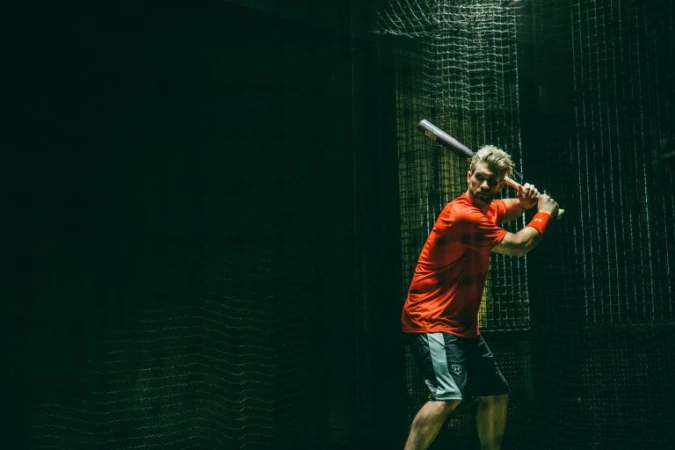 Adult man in athletic wear holds a baseball bat indoors within a netted batting cage with dark, dramatic lighting creating a sporty scene.
