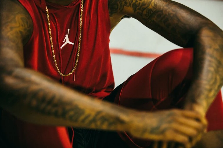 Torso of a man with arm tattoos, wearing a red hooded tank top with white logo and gold necklace, sitting indoors near a white wall.