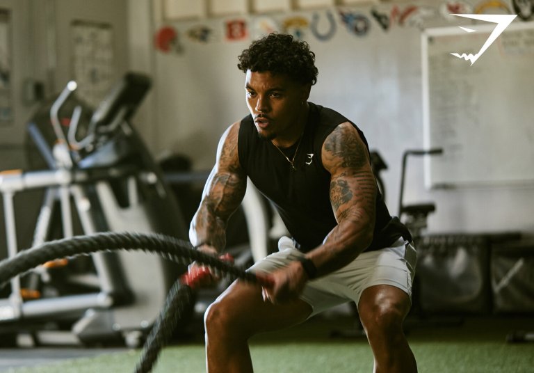 Athletic African-American man works out with battle ropes at indoor gym. He is wearing dark tank top and light-colored athletic shorts.