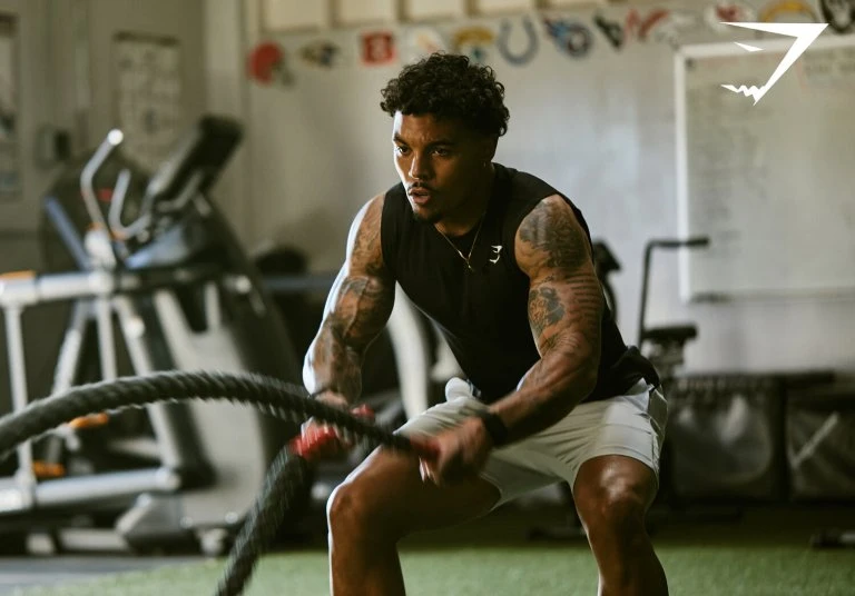 Athletic African-American man works out with battle ropes at indoor gym. He is wearing dark tank top and light-colored athletic shorts.
