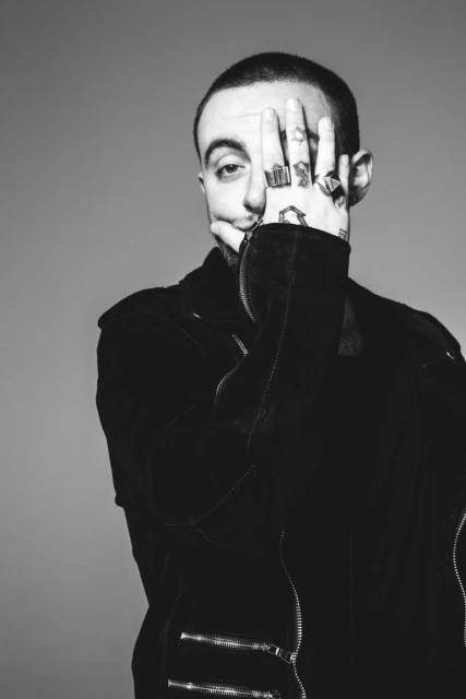 Black and white portrait of a fair-skinned adult male with rings covering one eye, wearing a black jacket, against a gray background.