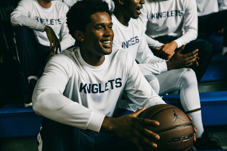 A group of male athletes sit on blue bleachers; one holds a brown basketball and smiles, wearing a white shirt and navy blue bottoms.
