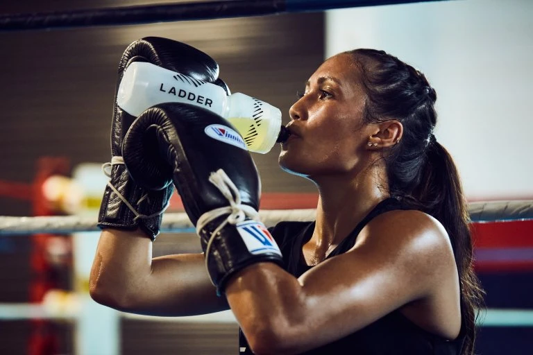 Woman drinks water from a white sports bottle in a boxing ring. She's wearing boxing gloves and a black tank top.