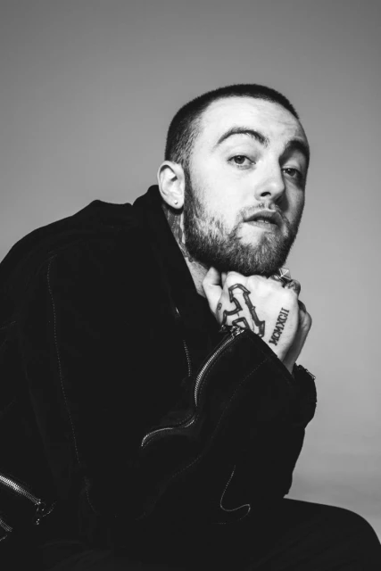 Black-and-white portrait of a man with a beard, resting his chin on his hands, looking at the camera, wearing a dark jacket.