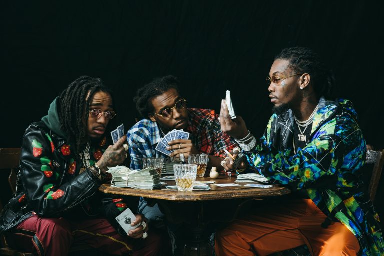 Three stylish men play cards at a table with cash, drinks, and serious expressions in a low-lit indoor setting, showcasing fashion and gaming.