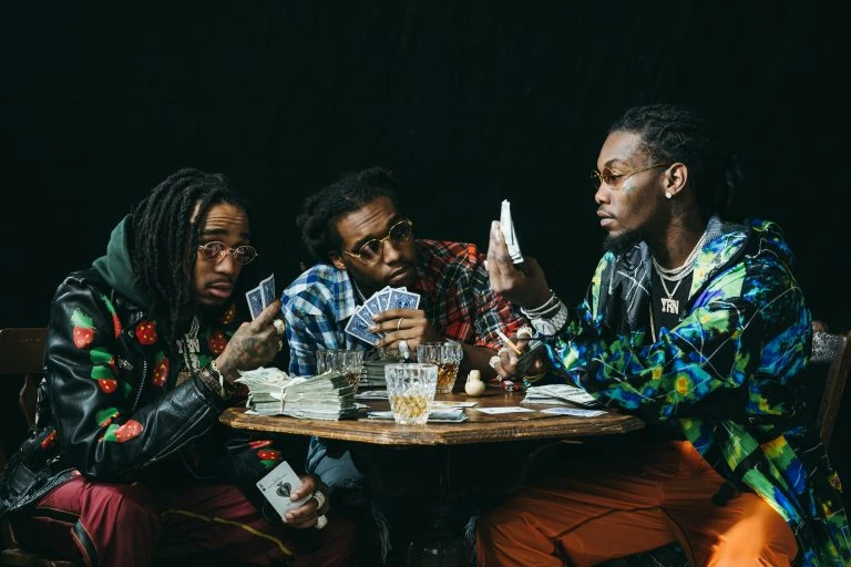 Three stylish men play cards at a table with cash, drinks, and serious expressions in a low-lit indoor setting, showcasing fashion and gaming.