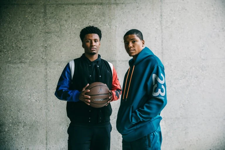 Two African American men in athletic jackets pose indoors with a basketball against a textured grey wall in a medium shot.
