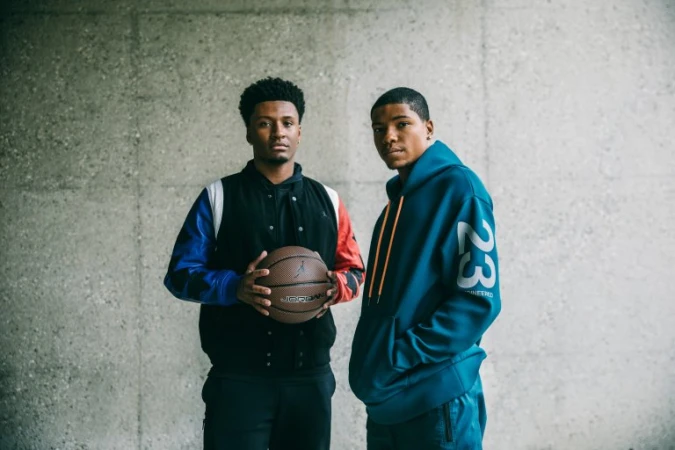 Two black men standing against a textured concrete wall, holding a basketball and wearing sportswear in Los Angeles.