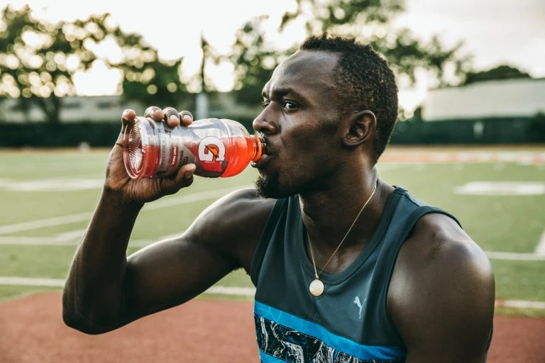 Adult male athlete, wearing a Puma tank top and a gold chain, drinks Gatorade on a running track on a sunny day outdoors.