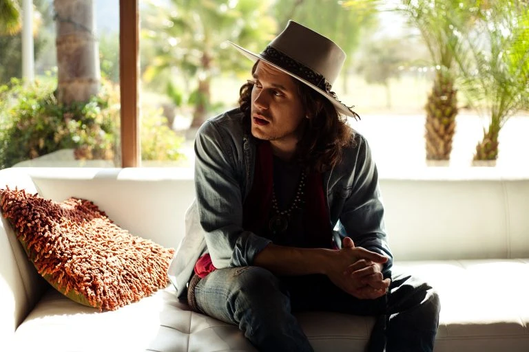 Man in hat sits on white couch in sunlit room with palm trees outside window, creating relaxed, stylish celebrity portrait.