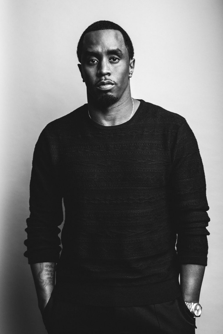 Black and white portrait of Sean Combs (P Diddy) in a dark sweater, hands in pockets, wearing a wristwatch against a plain studio background.