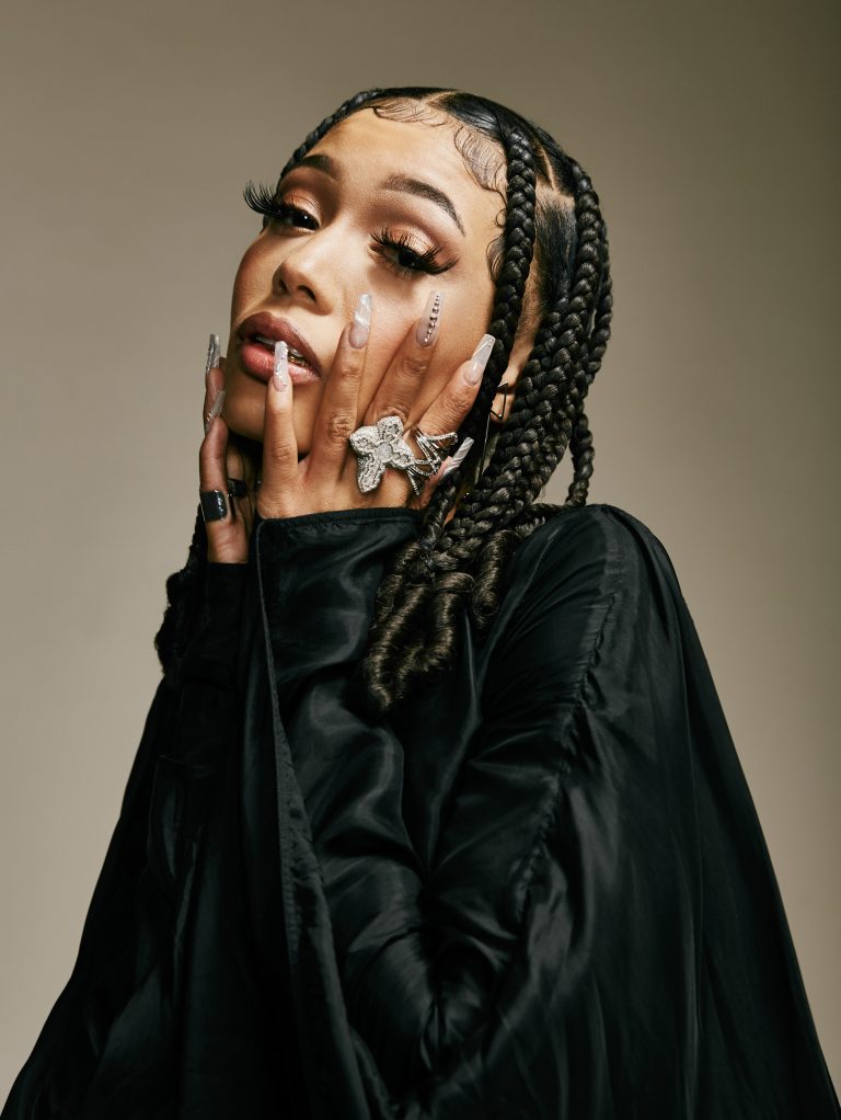 Portrait of woman with braids, butterfly ring, and black jacket posing in studio with neutral background and soft lighting.