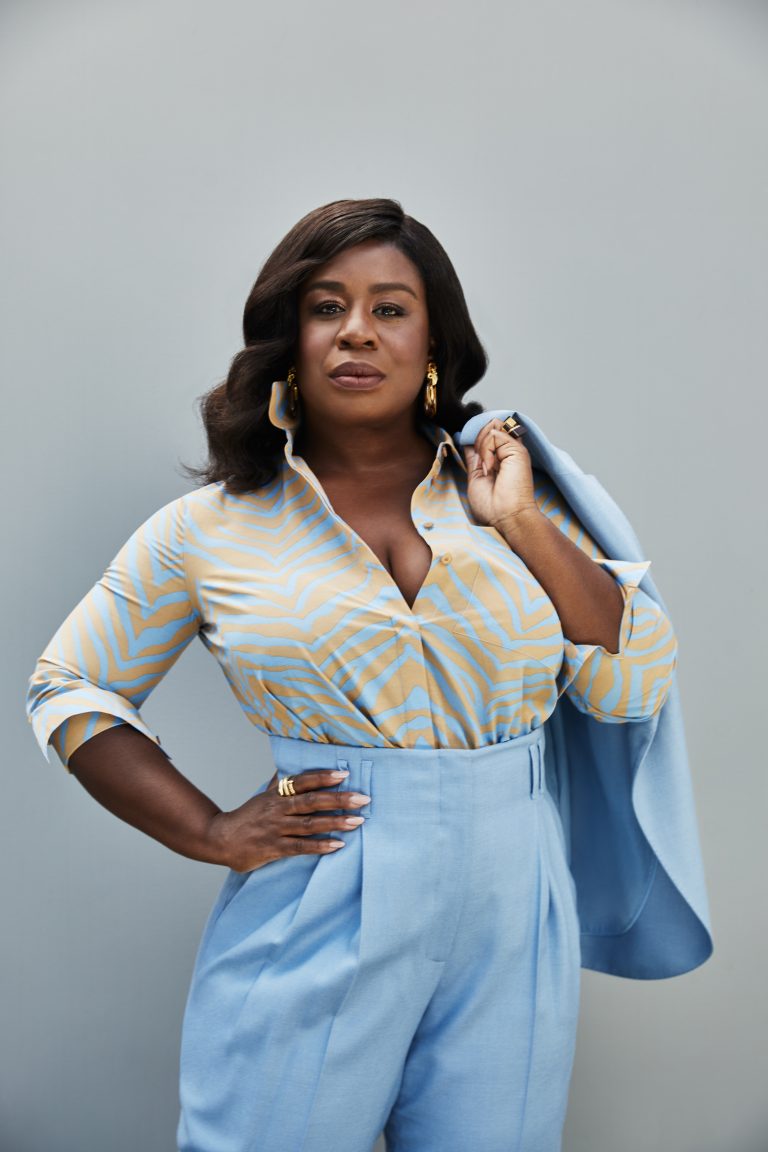 Portrait of a Black woman in a patterned blouse and high-waisted blue trousers holding a matching jacket over her shoulder.