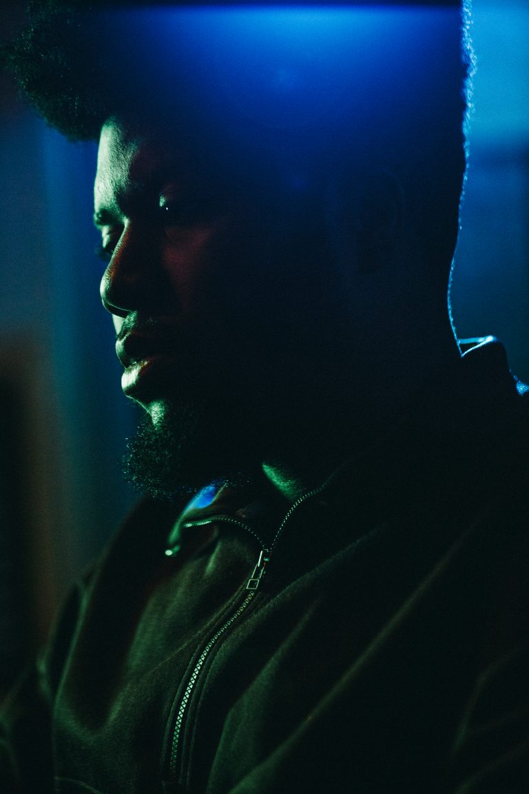 Low-angle portrait of a dark-skinned adult male in a hooded jacket, showcasing his face and torso against a blurred, dark, blue-lit backdrop.