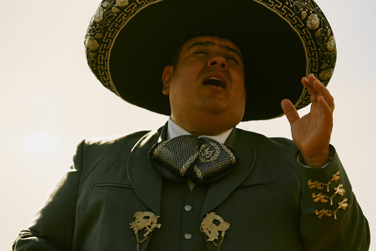 Adult male mariachi singer in traditional sombrero and charro suit performs outdoors against a bright sky, his hand raised as if conducting.
