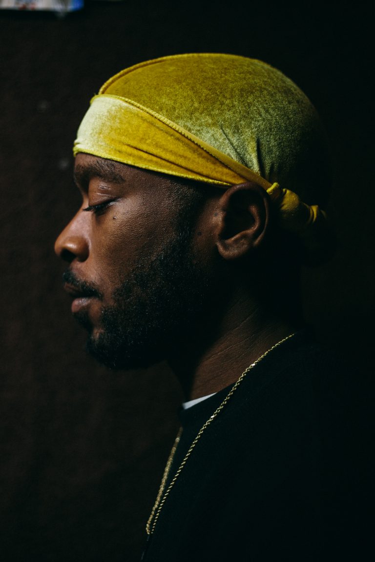 Close-up portrait of a Black man in profile wearing a yellow durag and gold chain against a dark backdrop.