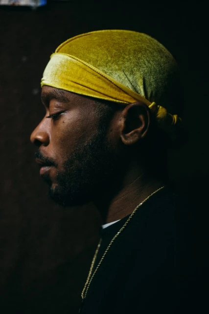 Close-up portrait of a Black man in profile wearing a yellow durag and gold chain against a dark backdrop.