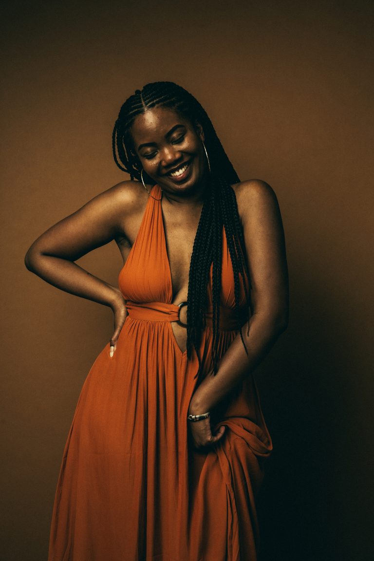 Smiling Black woman in a burnt orange plunge maxi dress with braided hair against a warm brown backdrop