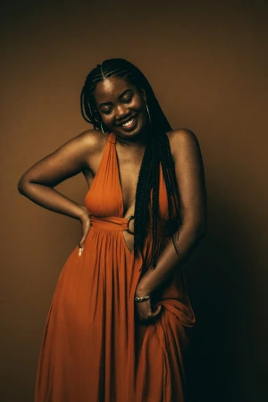 Smiling Black woman in a burnt orange plunge maxi dress with braided hair against a warm brown backdrop