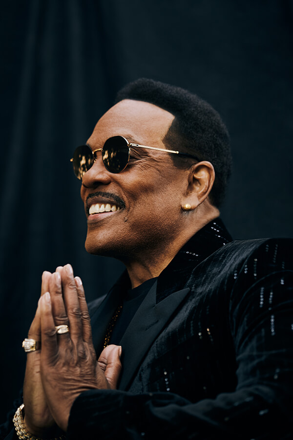Smiling African-American man wearing sunglasses and a velvet jacket, looking upwards with his hands clasped against a dark background.