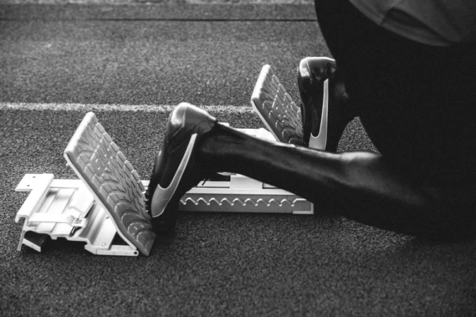 Black and white photo of an athlete's legs and feet positioned in the starting blocks on a textured running track surface.